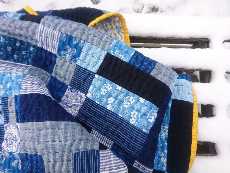 a blue rail fence quilt with bright yellow binding is crumpled on a snowy bench